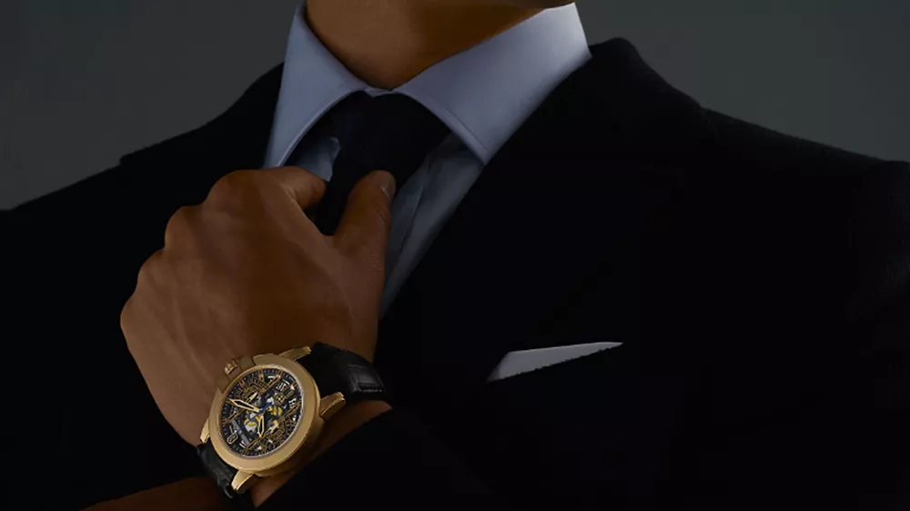 ساعت طلا مردانه امگا با بند چرمی مشکی در دست چپ مردی با کت و شلوار مشکی