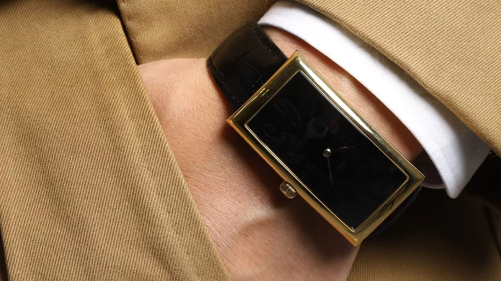 ساعت Tissot با قاب طلایی و بند چرمی مشکی در دست مردی با کت و شلوار قهوهای